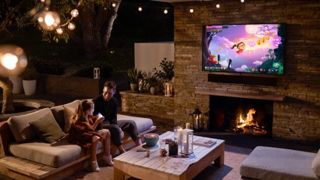 Father and daughter relaxing on an outdoor patio at night, watching a video game on a wall-mounted TV above a lit fireplace, with cozy seating, string lights, and a wooden coffee table in front of them.