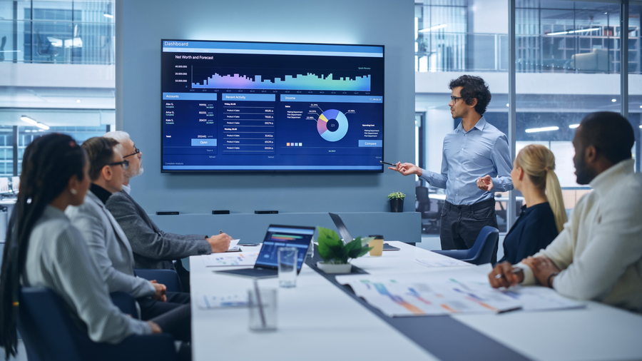 Business meeting in a modern office where a man presents financial data on a large wall-mounted screen to a group of five colleagues seated at a conference table with laptops, charts, and documents.