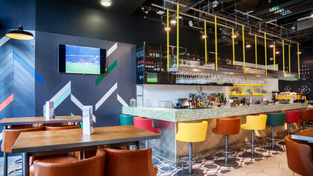 Modern restaurant bar with mounted TV, clean AV display, pendant lighting, and tabletop and bar seating.